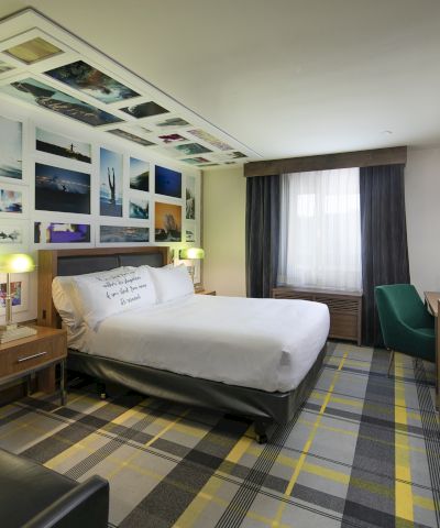 A modern hotel room with a bed, desk, TV, and a wall of colorful framed photos, plus curtains and lamps.