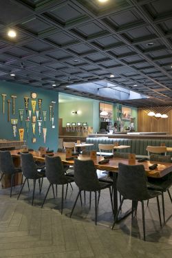 A modern indoor lounge/restaurant with tables, green booths, a bar counter, sports banners, and a lit paneled ceiling.