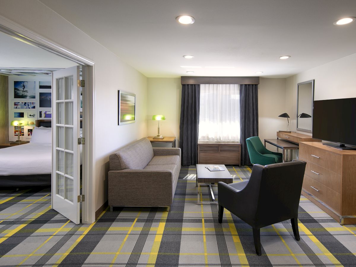 A modern hotel room with a sofa, desk, flat-screen TV, bedside lamps, and curtains, plus a striped carpeted floor.