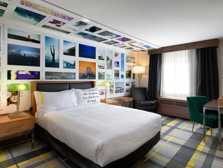 A stylish hotel room with a gallery wall of photos, a comfy bed, lamps, a desk, and a window with curtains.
