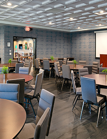A modern conference room with round tables, chairs, a bar area, and a projection screen under bright ceiling lights.